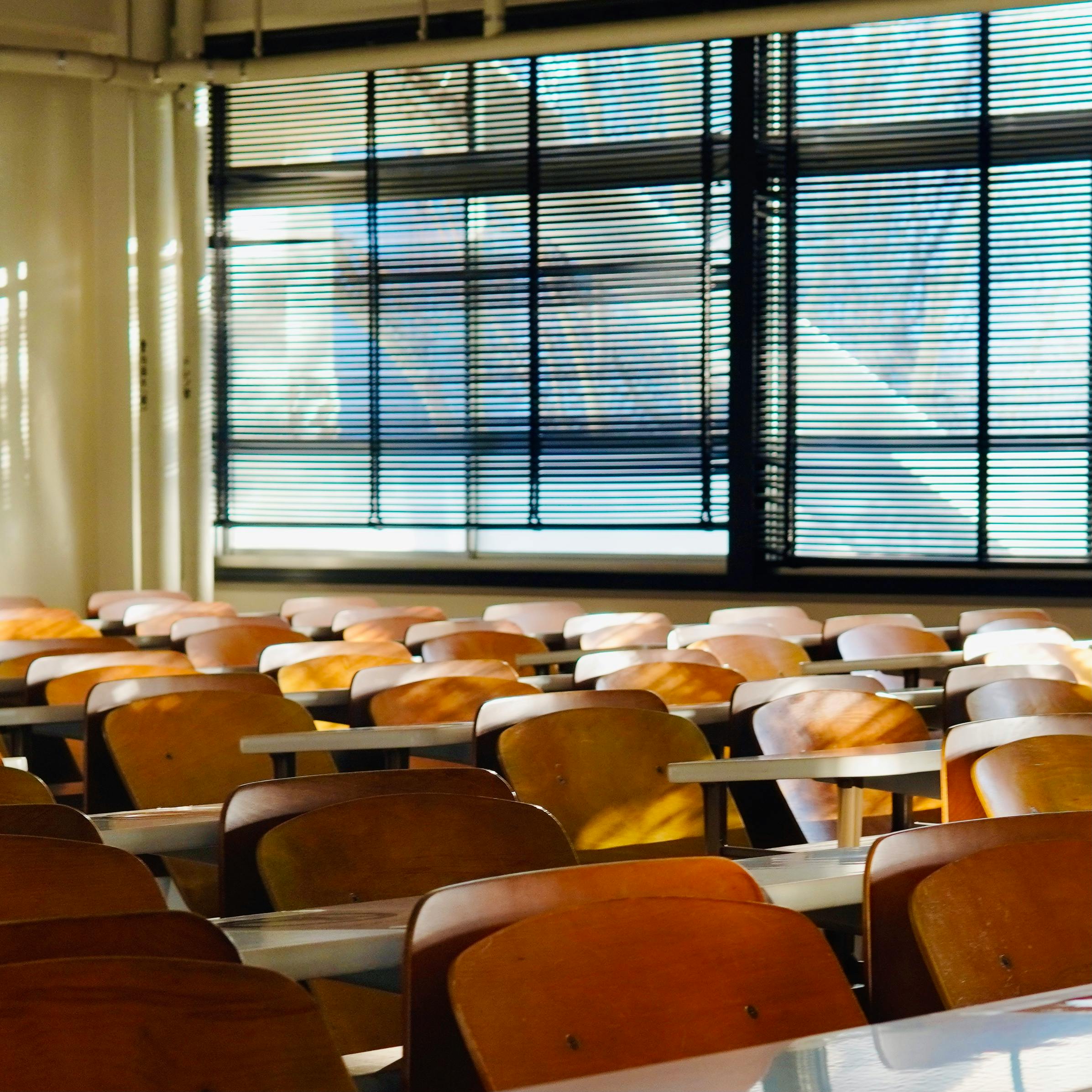 Sunlit empty classroom with wooden chairs and window blinds casting shadows across desks.